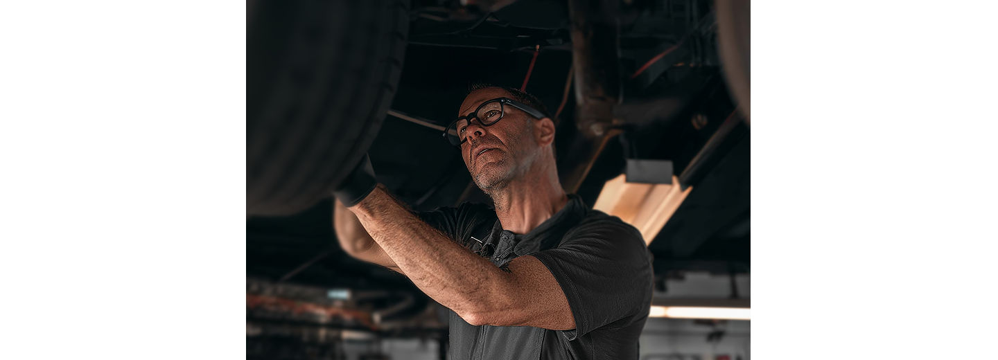 Automotive technician inspecting the underside of a vehicle raised on a lift inside a repair shop.