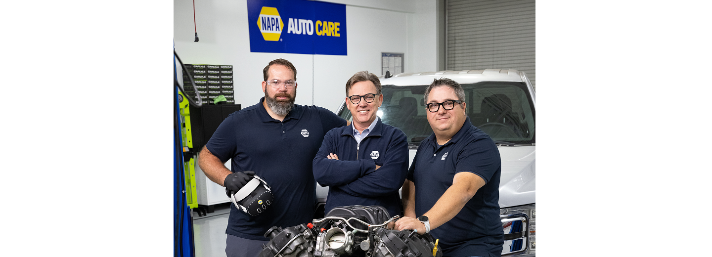 Three NAPA AutoCare representatives standing in an automotive training facility, with one holding a virtual reality headset and a vehicle in the background.